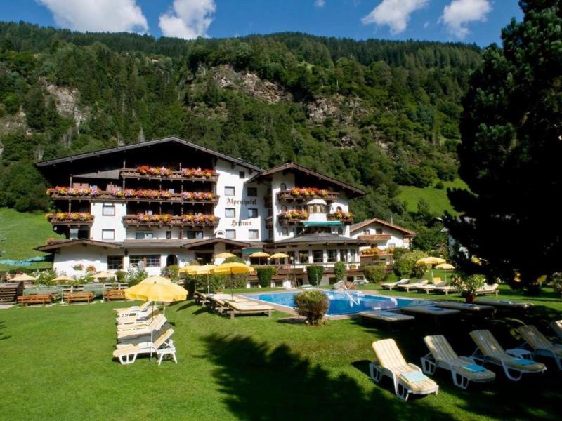 Großes Hotelgebäude mit Balkonreihen, grüner Liegewiese, Pool und Bergen im Hintergrund unter blauem Himmel.