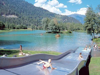 Kinder spielen auf einer großen Rutsche am See mit Wald und Bergen im Hintergrund.