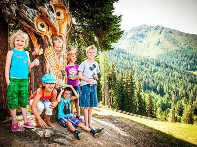 Gruppe fröhlicher Kinder vor einem Baum mit Pfauenfedern in einer bergigen Landschaft.