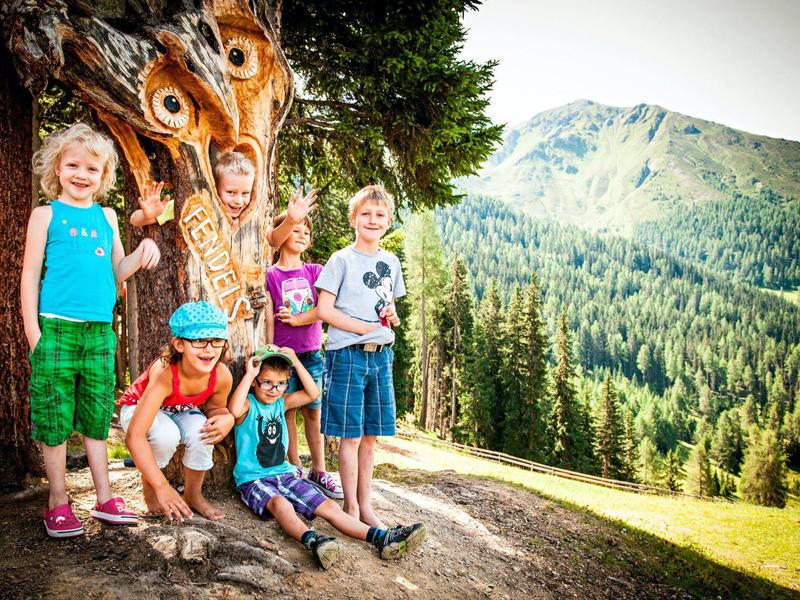 Gruppe fröhlicher Kinder vor einem Baum mit Pfauenfedern in einer bergigen Landschaft.