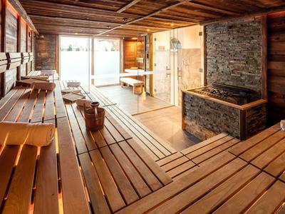 Helle Sauna mit Holzbänken und verputztem Steinofen, beleuchtet durch großes Fenster.