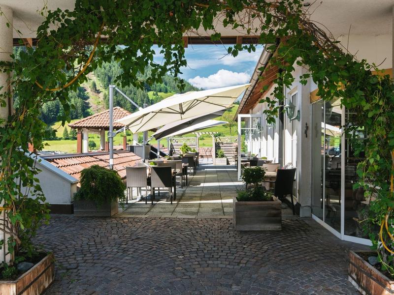 Gemütliche Terrasse mit Pflanzenbogen, Sonnenschirmen und Sitzgelegenheiten vor einem Hotelgebäude.