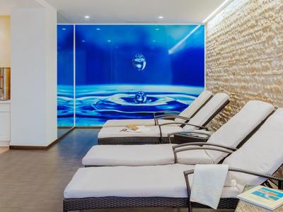 Drei Liegestühle mit weißen Polstern vor einer Wand mit Wassermotiv in einem modernen Wellnessbereich.