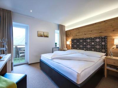 Helles Hotelzimmer mit Doppelbett, Holzwand, Sessel und Zugang zum Balkon.