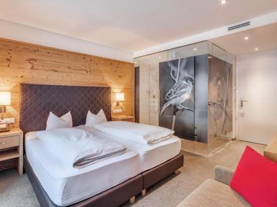 Modernes Hotelzimmer mit Doppelbett, Holzwand, Glastür zum Bad und rotem Kissen auf Sofa.