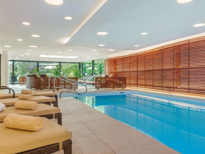 Innenpoolbereich mit Liegestühlen, Handtüchern und großen Fenstern mit Blick ins Grüne.