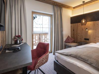 Gemütliches Hotelzimmer mit Holzelementen, Schreibtisch, rotem Stuhl und Balkon mit Blick nach draußen.