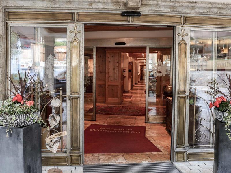 Eingang eines Hotels mit Glasschiebetüren und zwei großen Pflanzgefäßen mit roten Blumen.