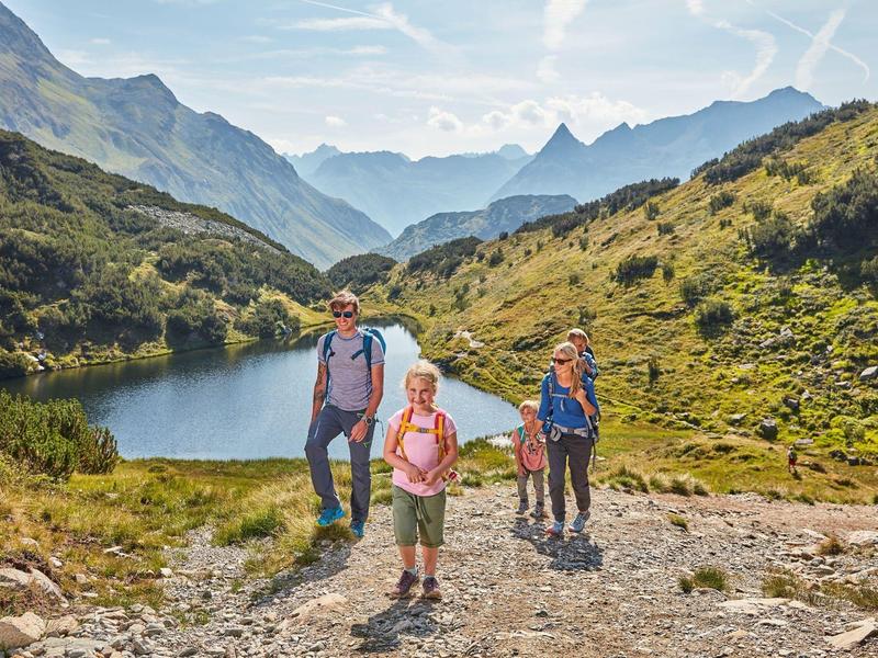 Drei Wanderer wandern auf einem felsigen Pfad in einer Berglandschaft mit See im Hintergrund.