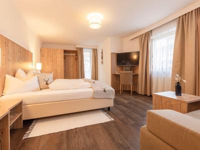 Helles Hotelzimmer mit Doppelbett, Holzmöbeln und Sitzbereich neben Fenster mit Vorhängen.