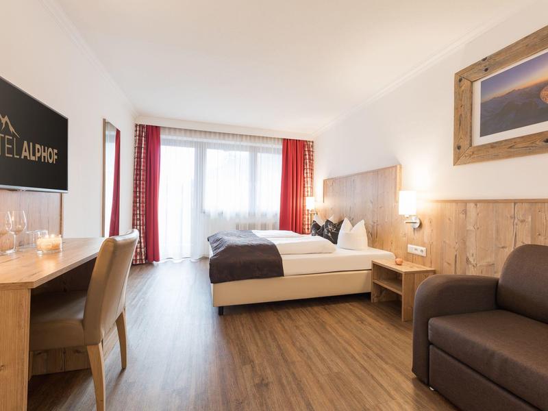 Helles Hotelzimmer mit Doppelbett, Holzfußboden, Schreibtisch und Sofa neben großem Fenster.