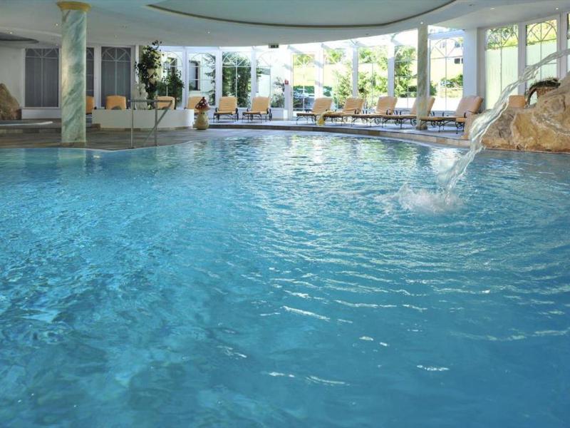 Hell beleuchteter Innenpool mit Wasserfall und Liegestühlen im Hintergrund.