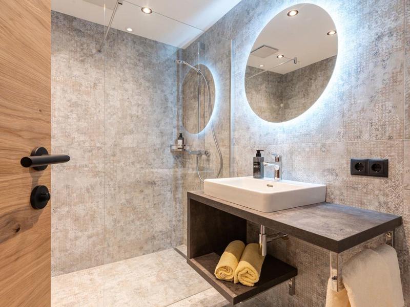 Modern bathroom with round illuminated mirror, sink, and wooden shelf with yellow towels.