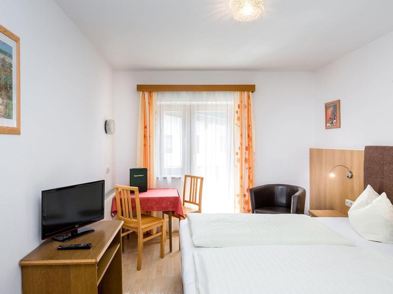 Bright hotel room with two single beds, TV, table with chairs, and balcony with curtains.