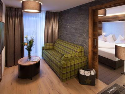 Modern eingerichtetes Hotelzimmer mit grün kariertem Sofa, Holzfußboden und angrenzendem Schlafzimmer.