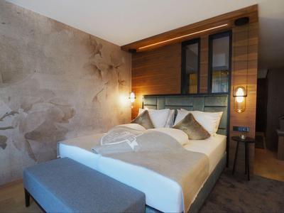 Modernes Hotelzimmer mit Doppelbett, Holzwand, Leselampen und einer Bank am Fußende.