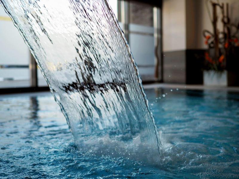 Nahaufnahme eines Wasserfalls in einem Hallenbad mit bläulichem Wasser und Fenster im Hintergrund