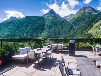 Gemütliche Terrasse mit Tischen und Stühlen vor bewaldeten Bergen unter blauem Himmel.