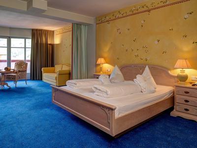 Geräumiges Hotelzimmer mit Doppelbett, blauem Teppich und Sitzbereich neben dem Fenster.