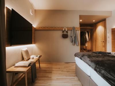 Modernes Hotelzimmer mit Holzboden, Bett mit dunkler Bettdecke und Wandhaken mit Taschen.