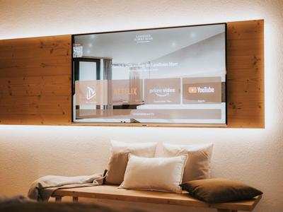Modern eingerichtetes Hotelzimmer mit Fernseher an der Wand und einem gemütlichen Sofa mit Kissen.