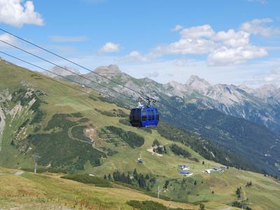 Blaue Seilbahn schwebt über grüne Berge mit Wanderwegen unter blauem Himmel.