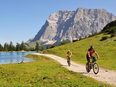 Zwei Radfahrer fahren auf einem Weg am See mit Bergen im Hintergrund.