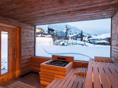Sauna mit Holzbänken und großen Fenstern, Blick auf schneebedeckte Berge und Häuser.