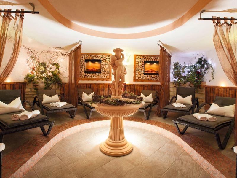 Eleganter Wellnessraum mit runder Fontaine, Holzmöbeln und warmen, goldenen Vorhängen.