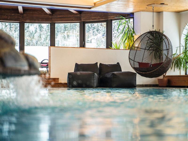 Innen-Pool mit hängendem Korbsessel, Pflanzen und Panoramafenstern, natürliche Holzelemente.