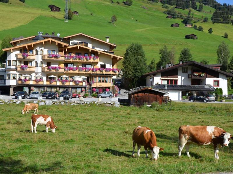 Grüne Bergwiese mit braunen Kühen vor mehrstöckigem Hotel und Hütten, im Hintergrund bewaldete Hügel.