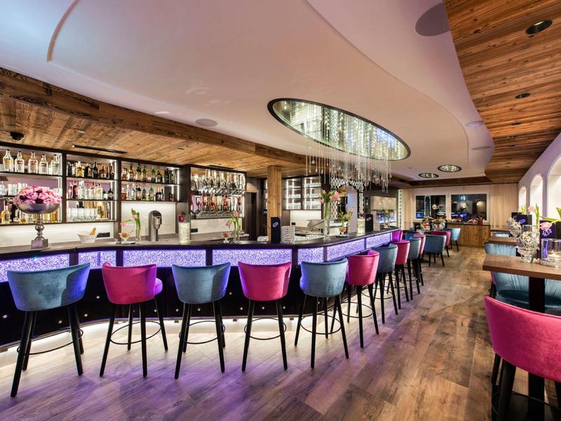 Moderner Barraum mit langer Theke, bunten Hockern und Holzdecke sowie warmem Holzfußboden.