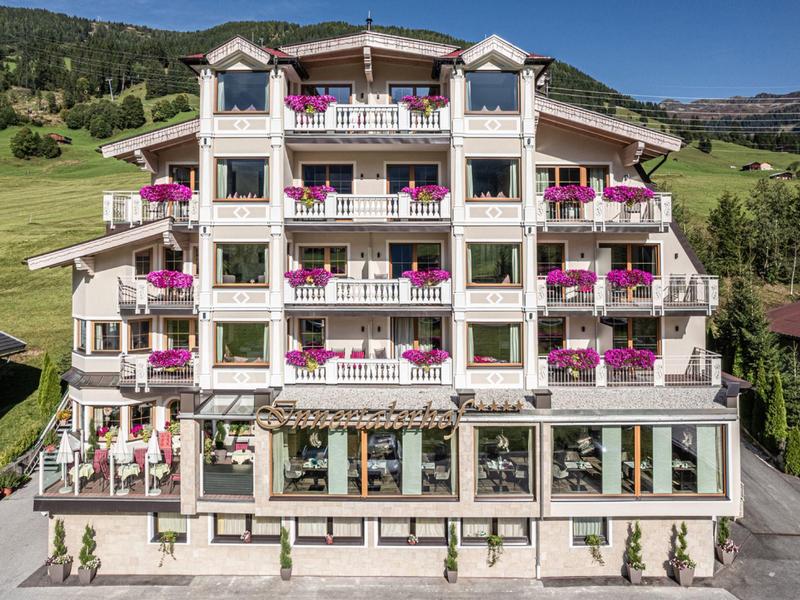 Großes weißes Hotel mit pinken Blumen auf Balkonen, Bergwiese im Hintergrund, klarer blauer Himmel.