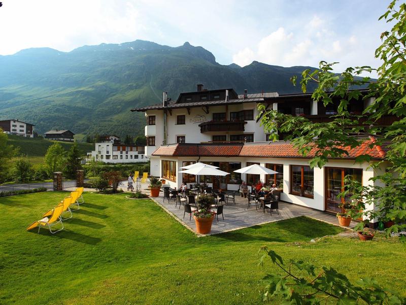 Hotel mit Terrasse und Sonnenschirmen vor grüner Berglandschaft unter bewölktem Himmel.