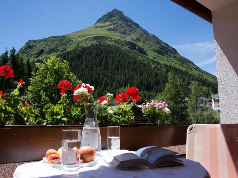 Bergblick vom Balkon mit Blumen, Tisch, Wasserflasche, Glas, Brötchen und offenem Buch.