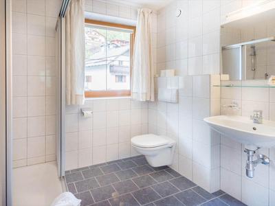 Helles Badezimmer mit weiß gefliesten Wänden, Fenster, Hänge-WC, Waschbecken und begehbarer Dusche.