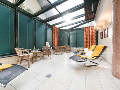 Hell gestalteter Wintergarten mit Liegestühlen und Rattanmöbeln, große Fensterelemente, weiße Fliesen.