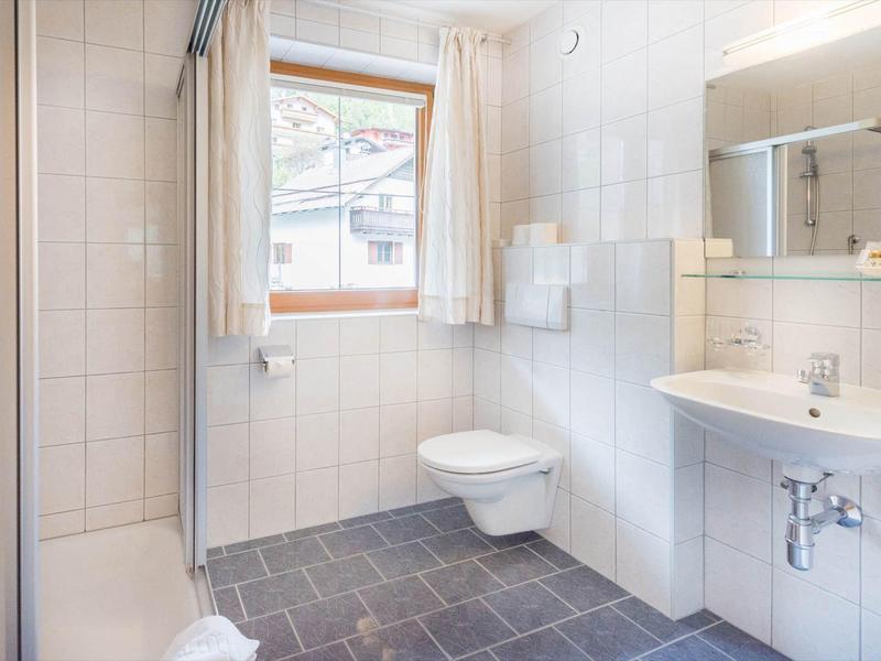 Helles Badezimmer mit weiß gefliesten Wänden, Fenster, Hänge-WC, Waschbecken und begehbarer Dusche.