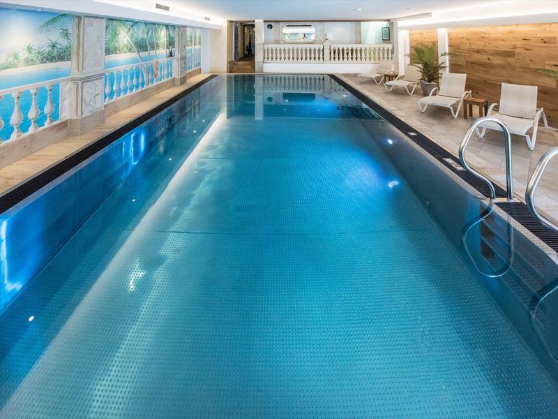 Innen-Schwimmbad mit blauem Wasser, weißen Liegestühlen und Wandmalereien mit Landschaftsmotiven.