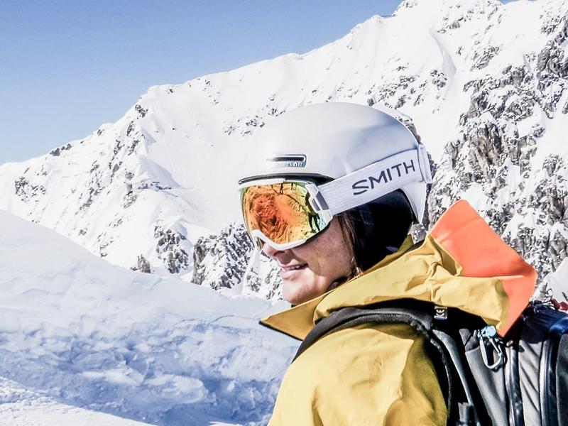 Mann in gelber Jacke mit Skihelm und Rucksack vor schneebedecktem Bergpanorama.
