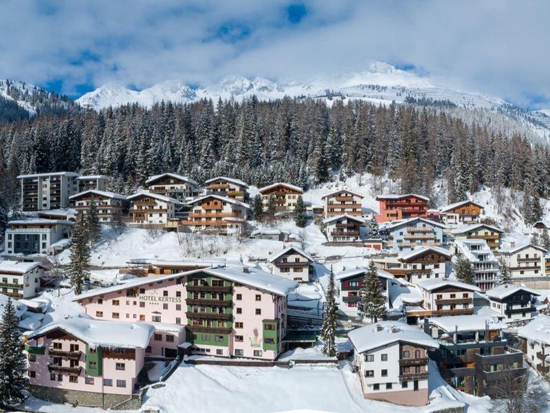 Winterliches Bergdorf mit schneebedeckten Häusern und Wäldern unter blauem Himmel mit Wolken.