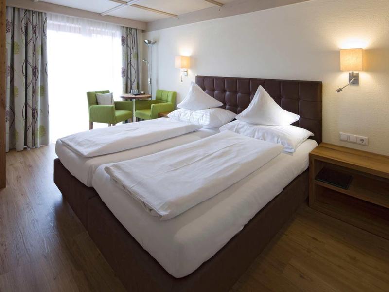 Gemütliches Hotelzimmer mit Doppelbett, grünen Stühlen, weitem Fenster und Holzboden.