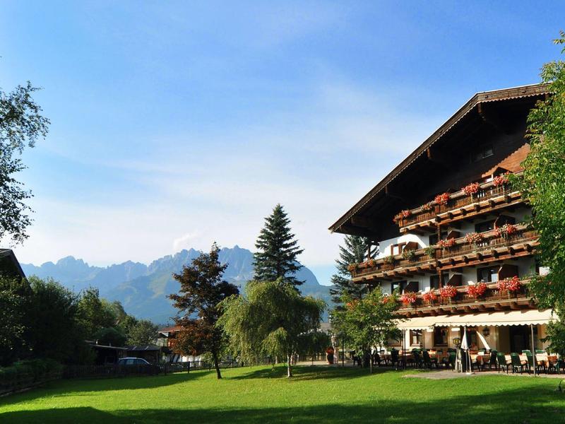 Großes traditionelles Holzgebäude mit Balkonen, umgeben von grünen Bäumen und Bergen im Hintergrund.