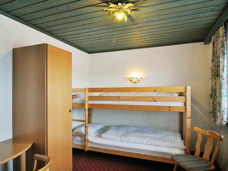 Kleines Zimmer mit Etagenbett, Holzschrank, Stühlen und Fenster mit Vorhängen.