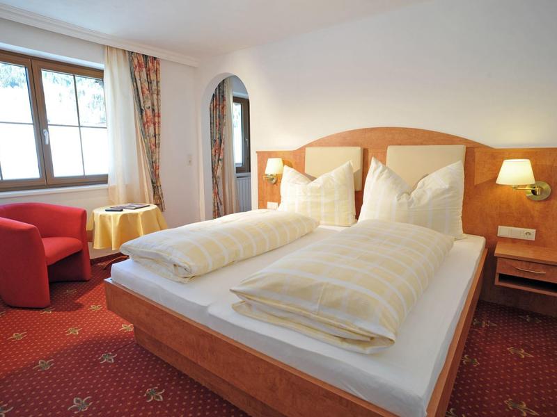 Gemütliches Schlafzimmer mit großem Bett, rotem Sessel, kleinem Tisch und Fenster mit Vorhängen.