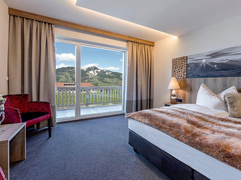 Modernes Schlafzimmer mit großem Bett, rotem Sitzbereich, Teppichboden und Balkon mit Bergblick.