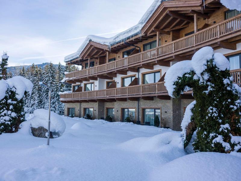 Schneebedecktes Holzhaus mit Balkon vor verschneiter Landschaft und Bäumen im Winter.
