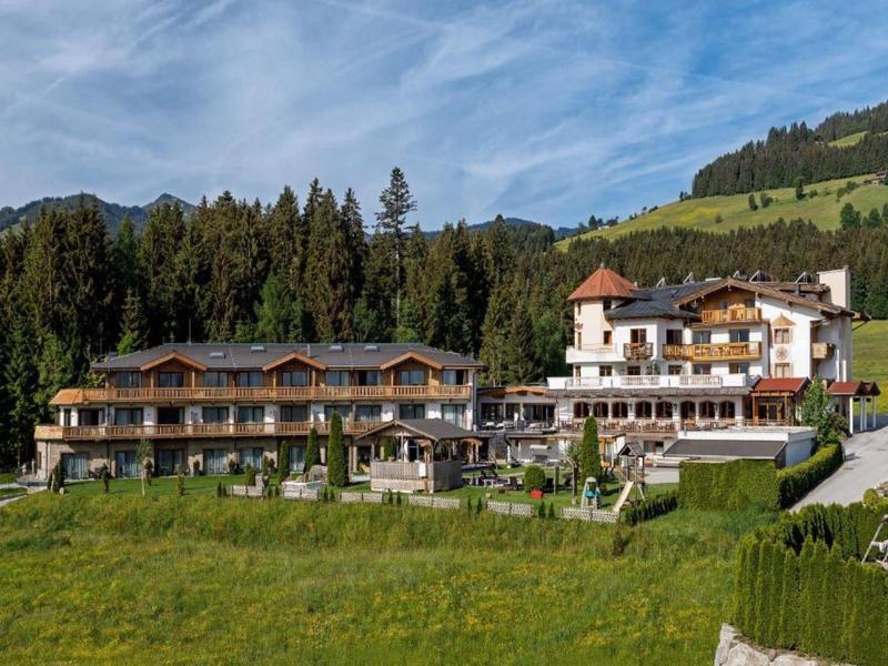 Großes Hotel mit mehreren Stockwerken, vor Wald und Bergen, umgeben von grüner Wiese unter blauem Himmel.