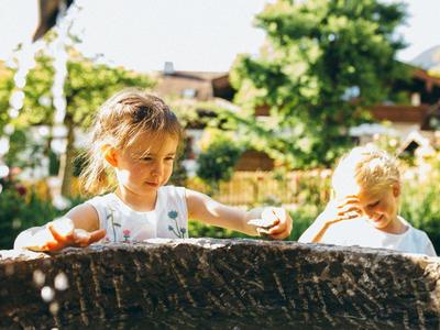 Zwei Kinder spielen draußen hinter einem grob strukturierten Holzzaun bei Sonnenschein.