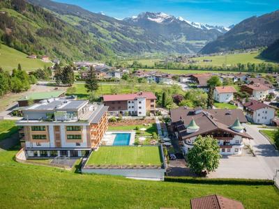 Blick auf ein Alpen-Dorf mit grünen Wiesen, Häusern, Bäumen und schneebedeckten Bergen im Hintergrund.